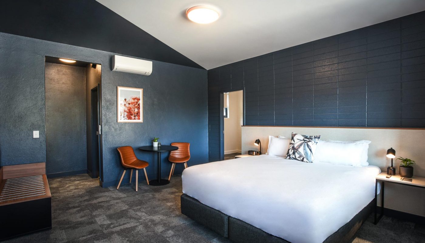 A large white bed sits in a modern room with dark paneled walls, two chairs, and a small desk.