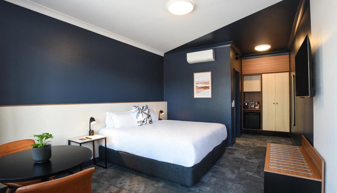 Bedroom with a white bed against navy walls, a small desk and plant, and an open doorway to a bathroom.