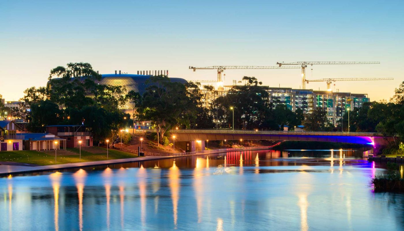 An illuminated bridge and city buildings at dusk reflect across a calm body of water.