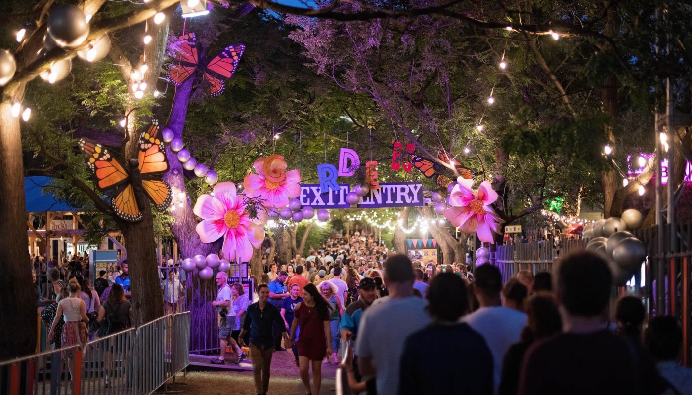 Crowds walk beneath a flower-decorated, brightly lit entrance arch at night, with string lights in the trees.