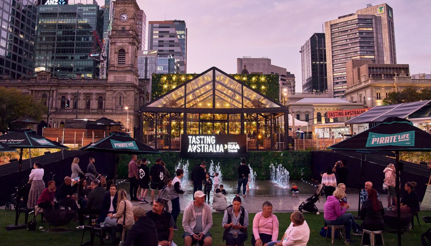 People gather at an outdoor event around a glass pavilion, with city buildings in the background at dusk.