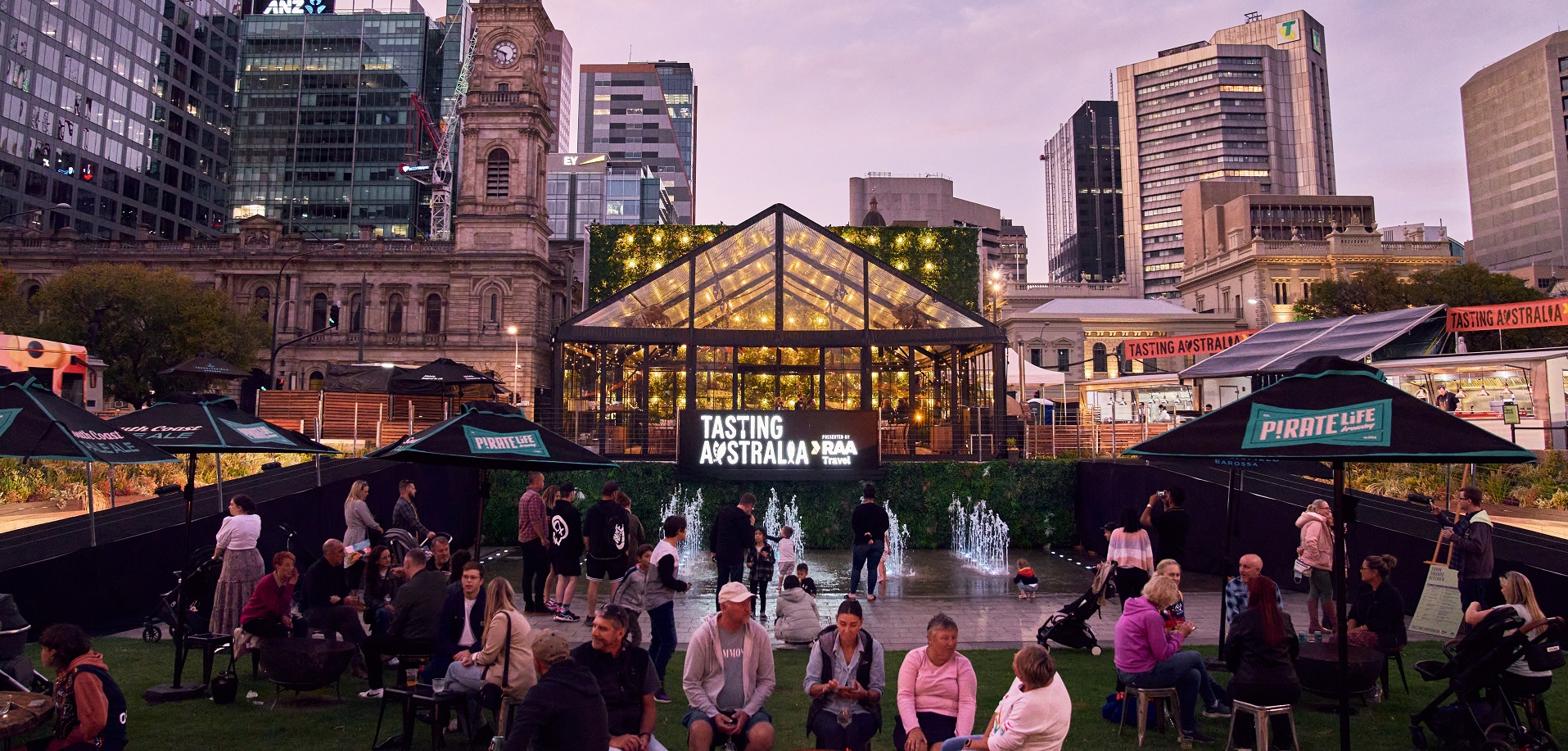 People gather at an outdoor event around a glass pavilion, with city buildings in the background at dusk.