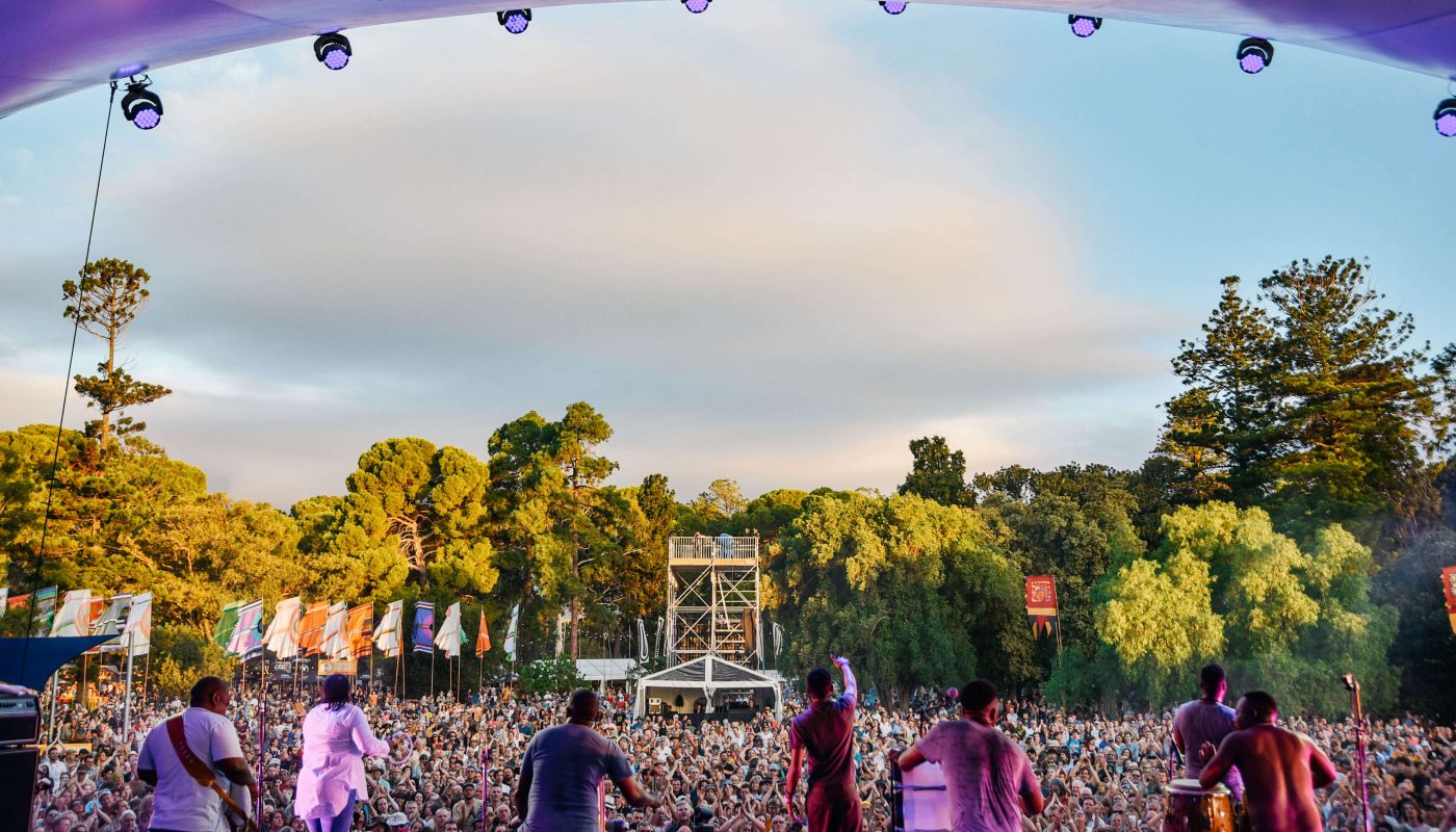 Musicians perform on an outdoor stage before a large crowd, with trees and a cloudy evening sky behind them.
