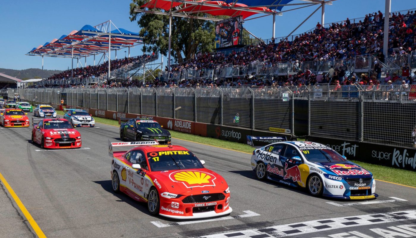 Several stock cars race down the track, with a red car leading past the start-finish line and packed grandstands beyond.