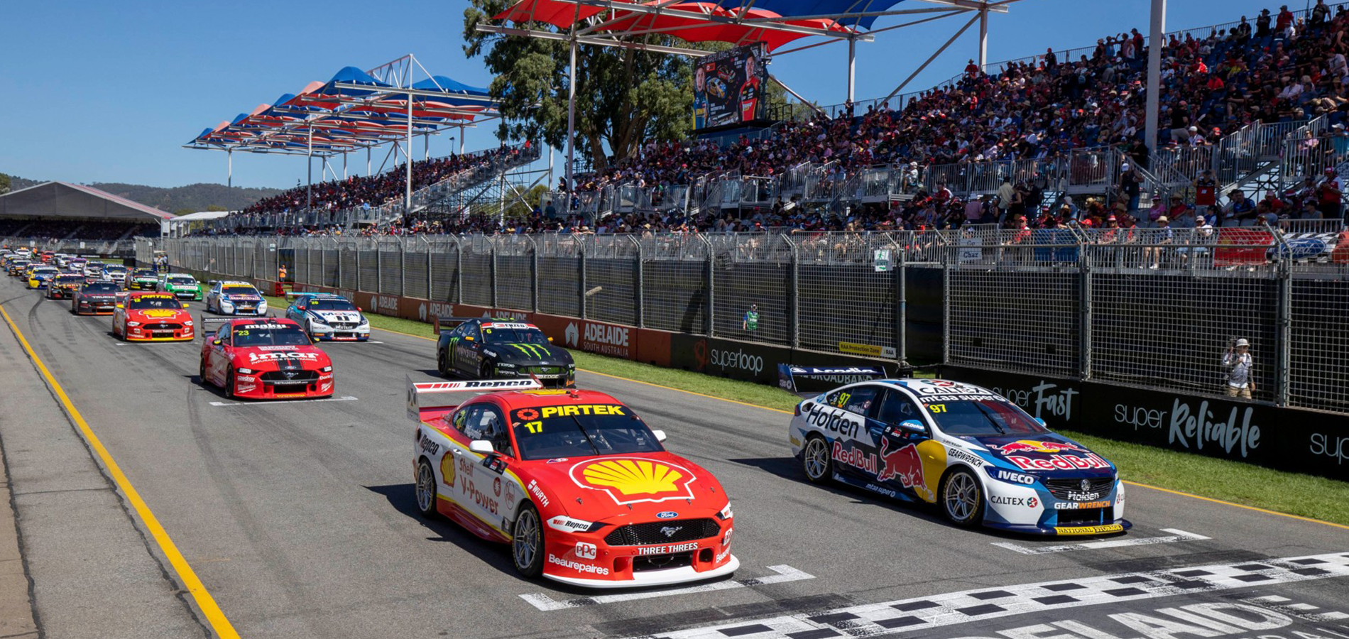 Several stock cars race down the track, with a red car leading past the start-finish line and packed grandstands beyond.