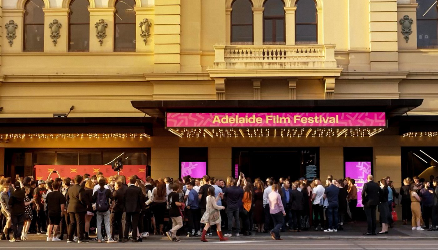 A crowd gathers outside a theater beneath a bright pink marquee.