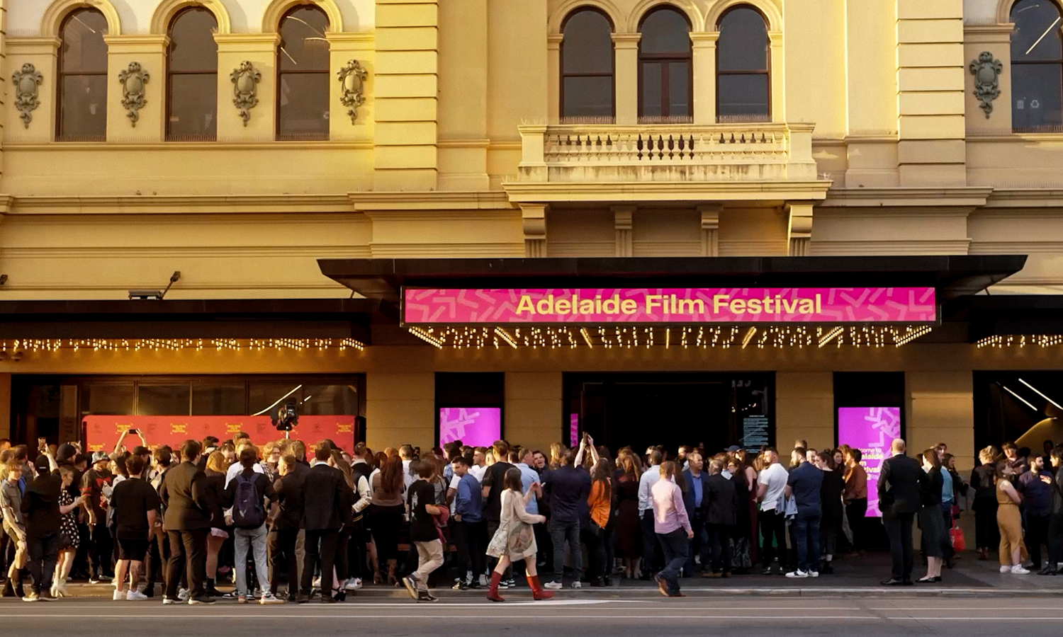 A crowd gathers outside a theater beneath a bright pink marquee.
