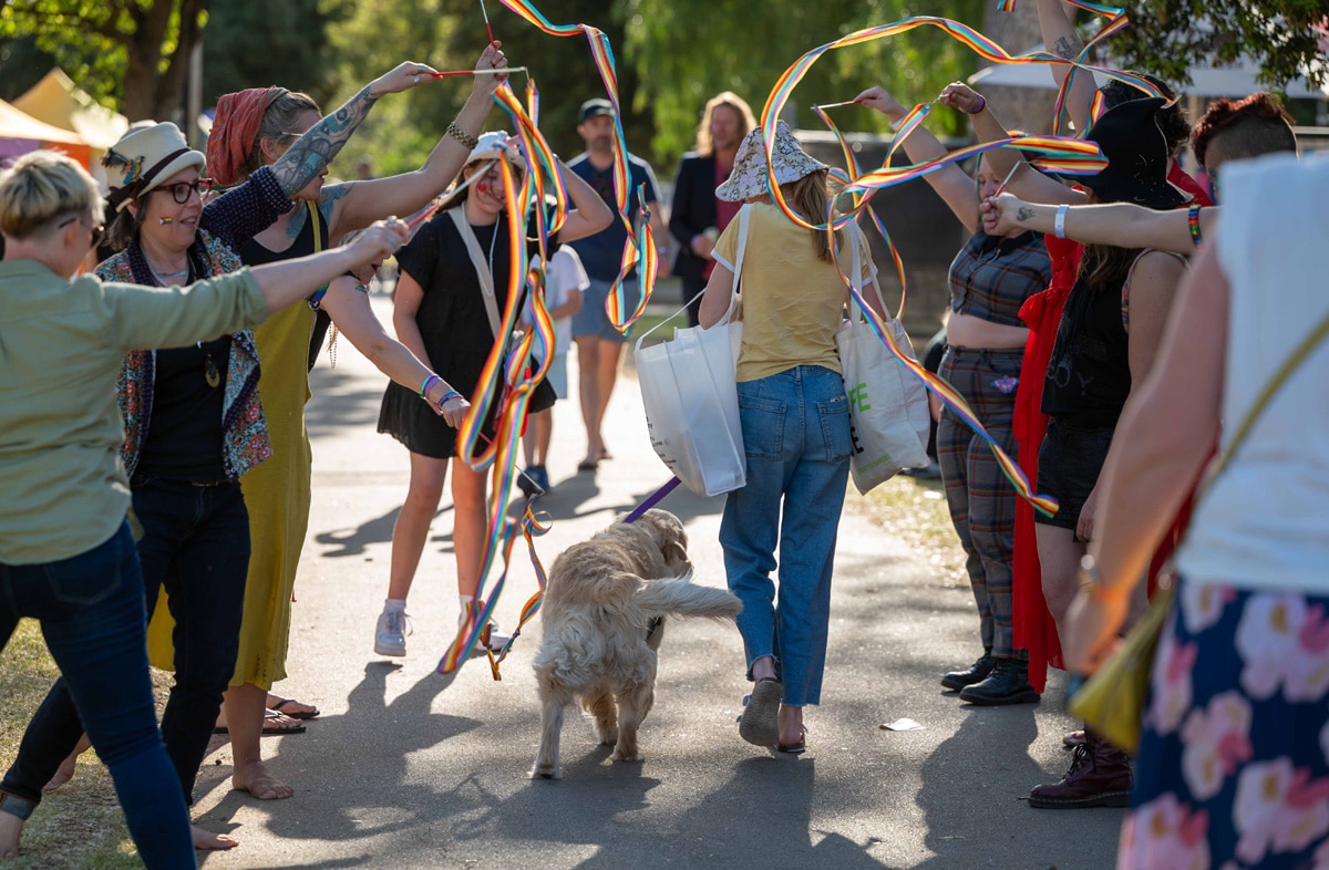 People hold colorful ribbons overhead as a small dog walks through the group outdoors.