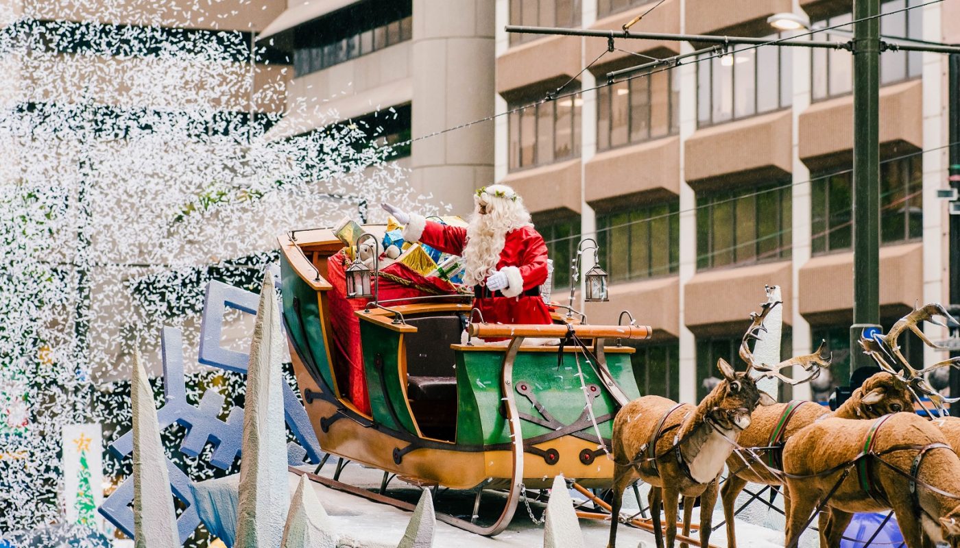 Santa rides in a decorated sleigh pulled by horses as confetti falls in front of a city building.