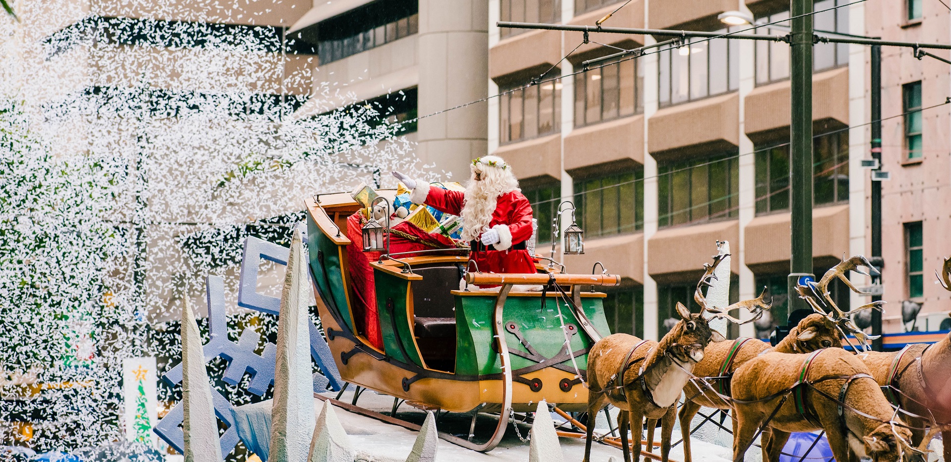 Santa rides in a decorated sleigh pulled by horses as confetti falls in front of a city building.