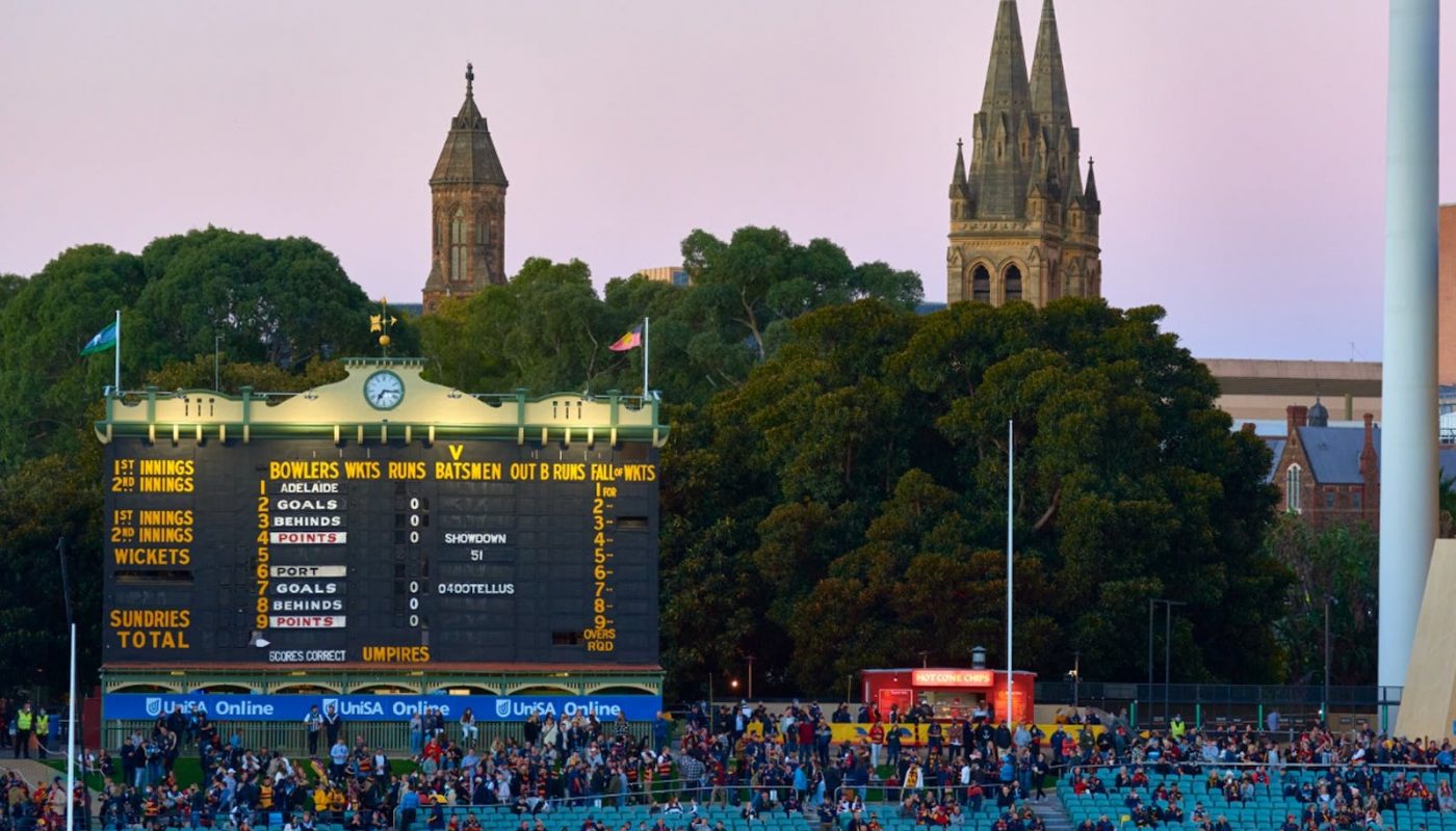 A crowded outdoor stadium at dusk, with a large scoreboard beneath trees and two church towers in the background.