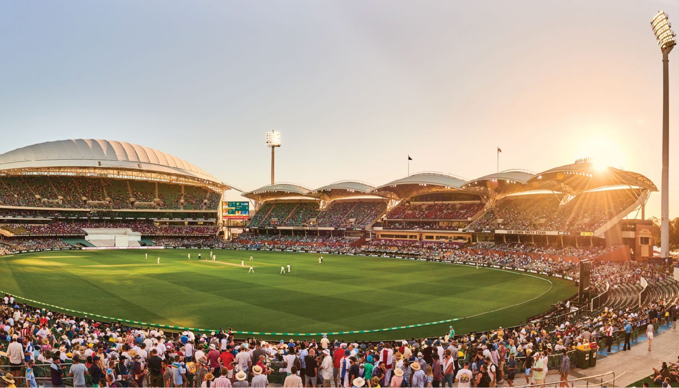 Crowded cricket stadium at sunset, with a green oval field surrounded by stands and floodlights.