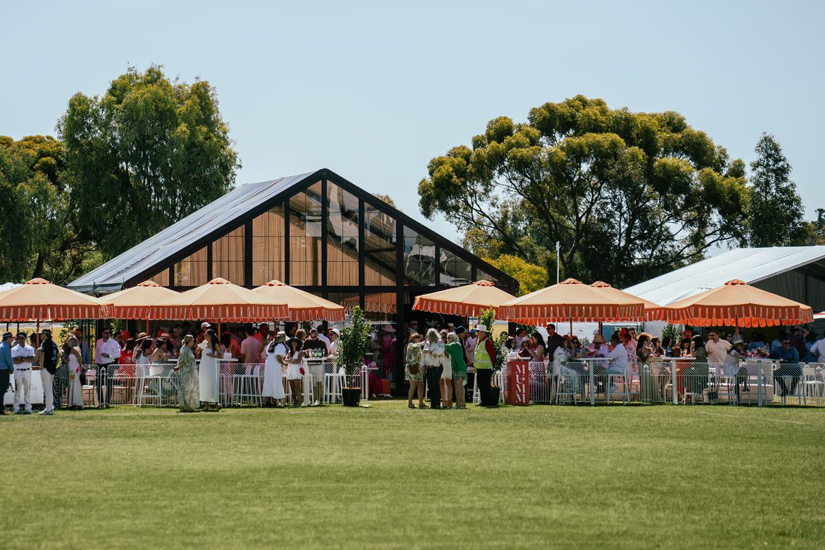 Guests gather beneath red-and-white umbrellas outside a glass pavilion, with a broad green lawn in the foreground.
