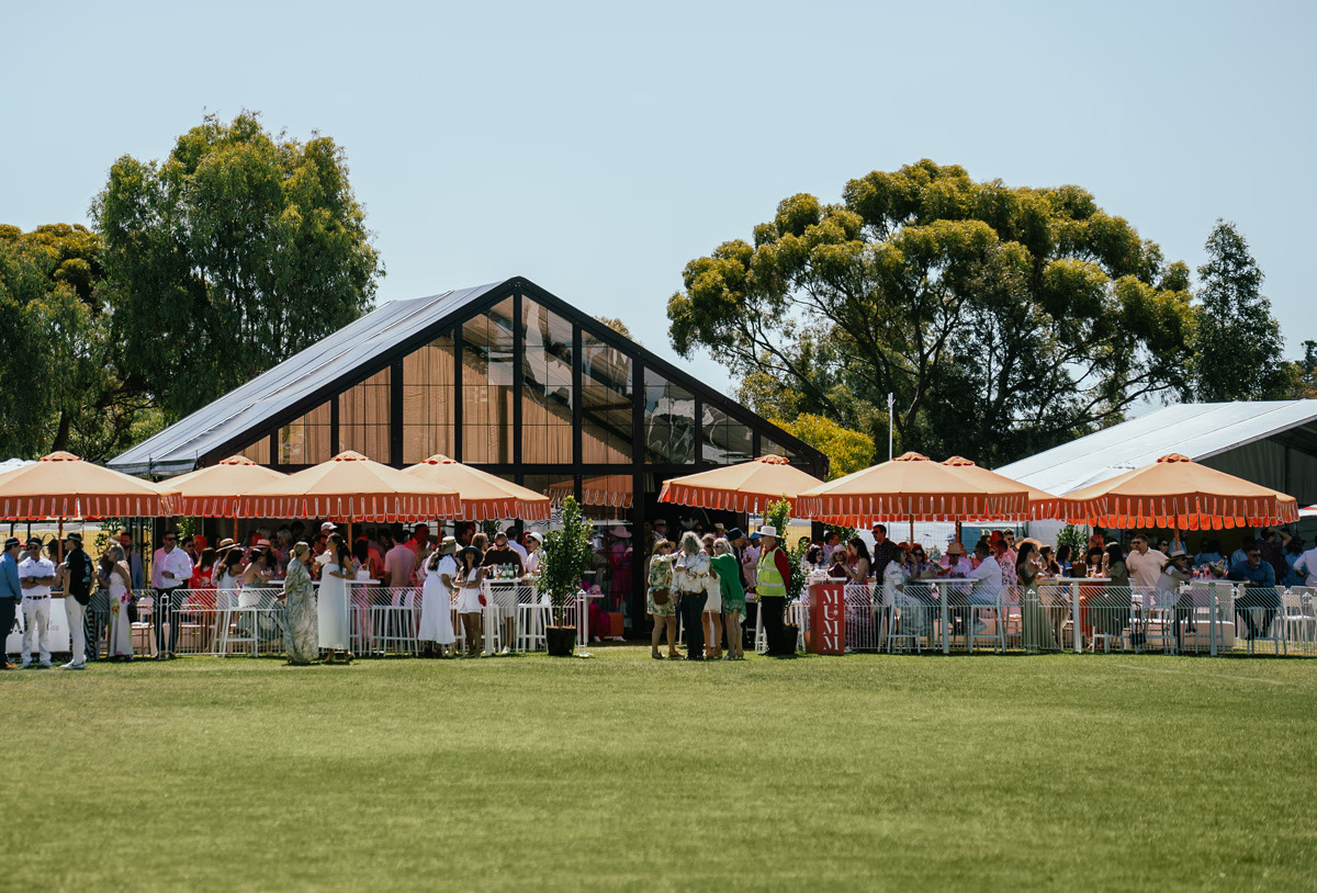 Guests gather beneath red-and-white umbrellas outside a glass pavilion, with a broad green lawn in the foreground.