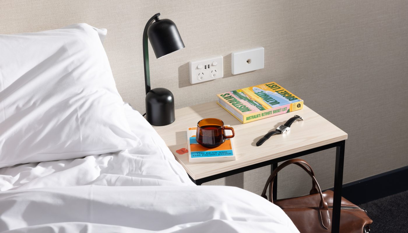 A white bed beside a table with a black lamp, wall outlets, a book, and small items; a brown bag sits underneath.