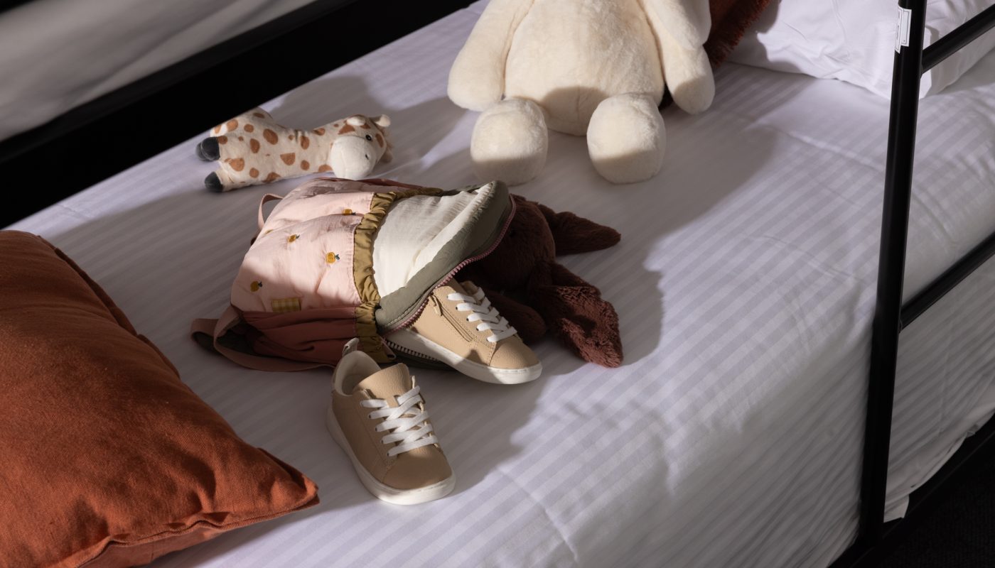 A striped bed holds a rust-colored pillow, folded clothes, small shoes, and plush toys.