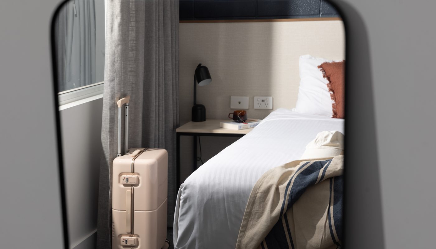 A rounded wall mirror reflects a bedroom with a neatly made bed, gray curtains, and a beige suitcase.