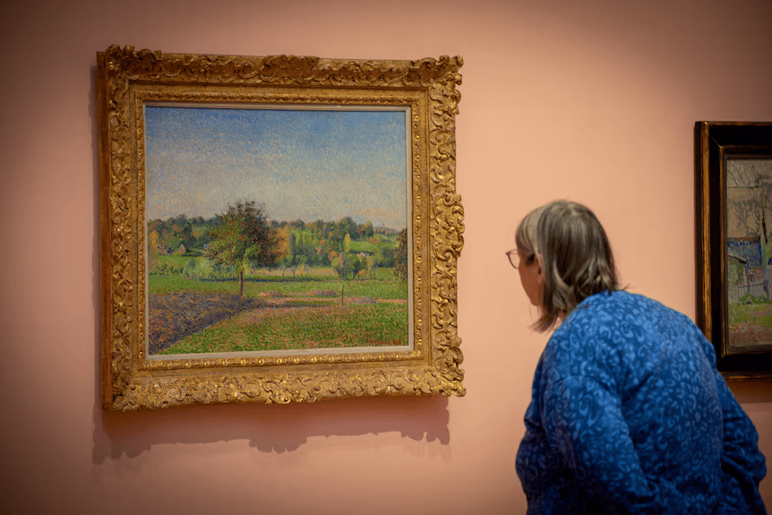 A person in a blue sweater looks at a framed landscape painting on a peach-colored gallery wall.