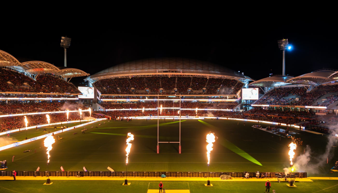 A brightly lit soccer stadium at night with a green field, packed stands, and flames shooting along the sidelines.