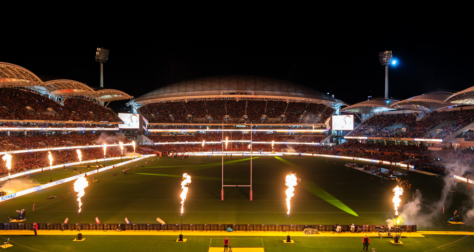 A brightly lit soccer stadium at night with a green field, packed stands, and flames shooting along the sidelines.